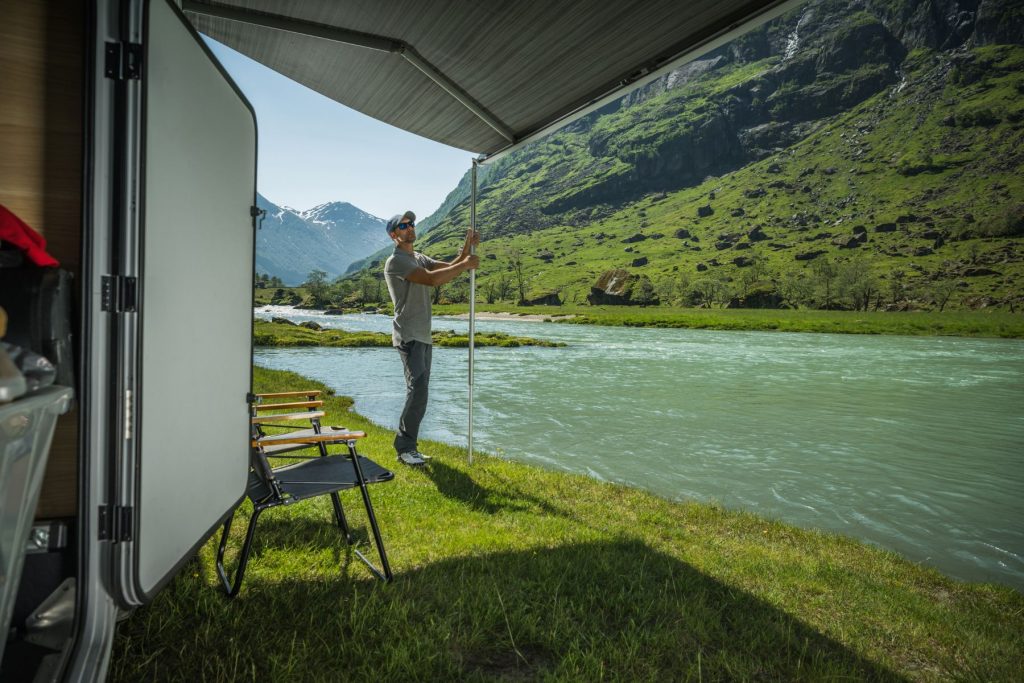 Man sets up the awning to his trailer which is parked next to a flowing stream.